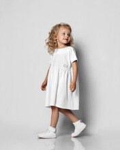 Little Blonde Curly Positive Princess Girl In White Casual Dress And Sneakers Standing Walking With Curly Hair Over Grey Wall
