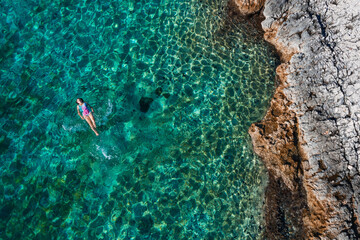 Wall Mural - Top aerial view of the young female floating on the back and relaxing on the warm turquoise Adriatic sea waves with rocky coastline. Carefree people vacation time concept image.