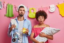 Family Portrait Of Father, Mother And Infant Pose At Home. Shocked Woman With Baby On Hands Stares At Camera, Displeased Tired Man Holds Newborn Items, Busy Nursing Daughter. Child Birth Concept