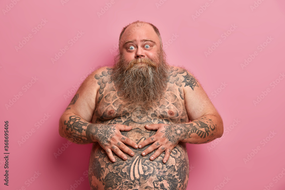 Bearded thick guy keeps hands on big tattoed belly, has bugged eyes, has thick beard, poses against pink background. Naked overweight adult man with large tummy, asks advice how to loose weight - obrazy, fototapety, plakaty 