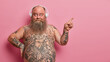 Cheerful self confident bearded man with big belly, tattooed body, listens pleasant music, wears headphones, points aside on blank space, advertises item on pink background. Promotion concept