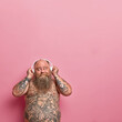 © WHstudio Leushin N - Vertical shot of thoughtful bearded man looks pleased above, keeps hands on headphones, thinks about something pleasant while listens music, has problem of obesity, tattooed naked body, stout tummy