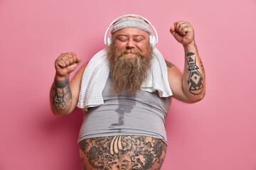 Wall Mural - Happy male fatso enjoys workout with music, raises hands and dances, has sweaty body, wears shirt and towel around neck, isolated on pink background. Chubby athlete happy to achieve great results