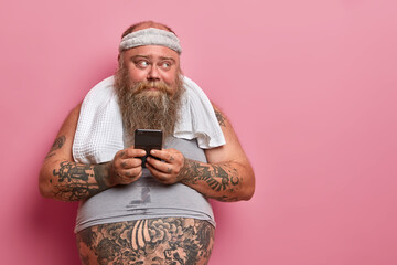 Wall Mural - Photo of overweight bearded man reads sms at smartphone, busy doing fitness at home, checks results in sport app how much calories he burnt, has tattooed belly sticking out from undersized t shirt