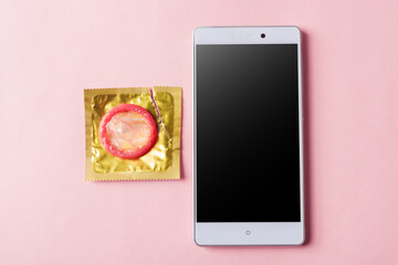 World sexual health or Aids day, Top view flat lay condom in wrapper pack and smart mobile phone blank screen, studio shot isolated on a pink background, watching porn content on mobile phone concept