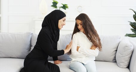 Wall Mural - Pretty smiled Arabian mother in black hijab talking with small teen daughter and calming her down. Muslim woman in headscarf having talk with little girl on couch at home and hugging. Mom's love hugs.