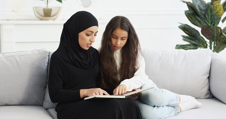 Wall Mural - Young beautiful Arabian woman in black hijab trading interesting story in book for pretty small teen girl. At home on couch. Muslim mother learning and educating little teenage daughter with textbook.