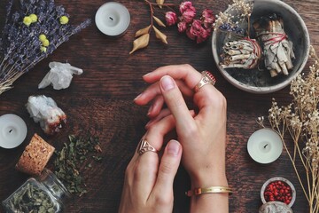 Witches hands on a table ready for spell work. Wiccan witch altar filled with sage smudge sticks, herbs, white candles. Female witch wearing vintage jewellery, placing her hands on dark wooden table