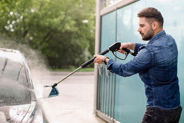 Wall Mural - Handsome bearded young man in jeans shirt, washing his new car manually with water high-pressure hose at outdoor self wash service. Car washing concept.