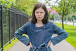 © Николай Батаев - An upset girl stands with her hands on her hips on the street against the background of the park behind the fence. Concept - bad mood, frustration and depression