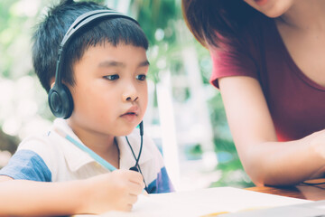 Wall Mural - Young asian mother and son using laptop computer for study and learning together at home, boy writing on notebook for homework and wearing headphone, teacher or mom support child, education concept.