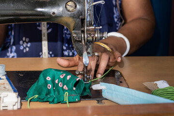 Indian woman working on old sewing machine, making homemade face masks against coronavirus or covid19 spreading, closeup detail on moving needle and fingers holding fabric	