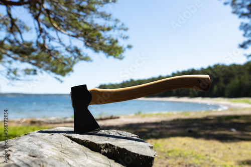 Axe ready for cutting timber. Axe in stump. Lumberjack axe in wood, chopping timber. Travel, adventure, camping gear, outdoors items. Background of water and a blue sunny sky.