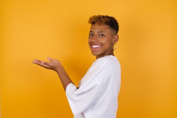 Young African American girl with afro short hair wearing white tshirt  pointing aside with hands open palms showing copy space, presenting advertisement smiling excited happy