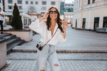 Stylish woman with camera looking up while walking down the street. Outdoor photo of carefree brunette girl in sunglasses smiling on city background.