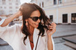 © Look! - Amazing brunette woman in trendy earrings smiling on city background. Wonderful tanned girl in sunglasses enjoying good day.