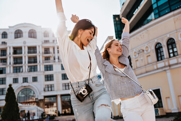 Wall Mural - Stunning female photographer dancing on the street with friend. Stylish girls having fun outdoor in sunny day.