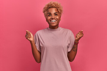 Wall Mural - Portrait indoors of a dark-skinned girl with a short haircut who finally got what she really wanted. Girl in casual clothes stands on a background of a pink wall and is sincerely happy.