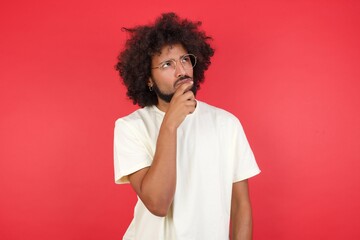 Shot of contemplative thoughtful Young man keeps hand under chin, looks thoughtfully upwards, dressed in casual clothes, poses over white background with free space for your text