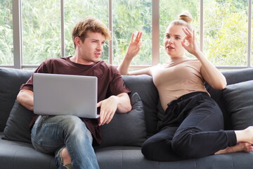 Young Caucasian couple sitting on sofa at home with computer and talking to each other. Couple lifestyle spending time together at home on their weekend concept