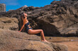© unai - Lifestyle, Caucasian brunette in a luxurious black swimsuit with sparkles enjoying the summer at sea. Portrait sitting on a rock with the sea behind