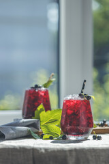 Two glass of cold ice black currant juice with ripe berries and green leaves on table in sunny room near window with garden outside.