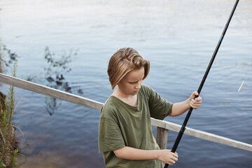 Wall Mural - Happy boy fishing from wooden placing, holding fishing rod in hands and being ready to cast line, blonde guy spending time near lake, wants to catch big fish.