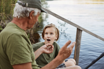 Wall Mural - Father and son fishing near river, dad telling about last fishing, showing size of catching fish, son listening with astonished facial expression and opened mouth.