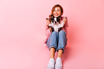 Wall Mural - Well-dressed girl playing with french bulldog. Indoor photo of beautiful red-haired woman expressing sincere emotions.