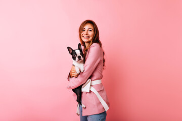 Wall Mural - Pleasant caucasian woman in pink attire embracing puppy. Glamorous white girl holding french bulldog with smile.