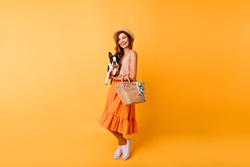 Wall Mural - Full-length portrait of romantic young lady with summer hat holding dog. Stunning red-haired girl in long skirt posing with french bulldog.