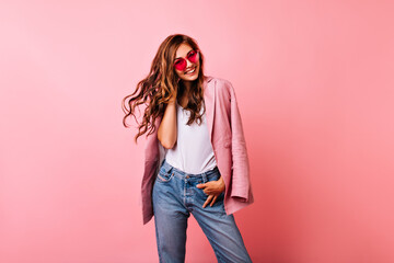 Wall Mural - Excited white girl in bright stylish glasses posing on pink background. Dreamy curly woman playing with her ginger hair and laughing.