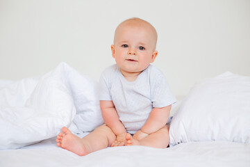 Cute baby boy 6 months smiling in a white bodysuit sitting on a bed on white bedding