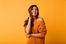 Adorable Woman In Orange Attire Touching Her Brown Wavy Hair. Laughing Blithesome Girl Posing On Yellow Background.
