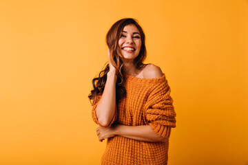 adorable woman in orange attire touching her brown wavy hair. laughing blithesome girl posing on yel