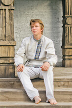 Young Man In Old Linen Clothes Is Sitting On The Steps Of The Porch Of A Wooden Terem