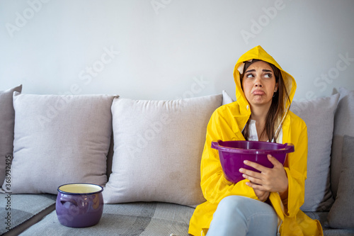 House Ceiling is Flowing - Woman in raincoat Holding Bucket While Water Droplets Leak From Ceiling. Shocked Woman Looks at the Ceiling While Collecting Water Which Leaks in the Living Room at Home.