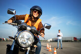 Female student with helmet taking motorcycle lessons and practicing ride. In background traffic cones and instructor with checklist rating and evaluating the ride. Motorcycle school of driving.