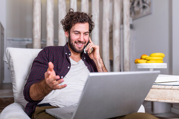 Wall Mural - Smiling handsome man talking on smartphone and using laptop computer while sitting at home. Freelance Career. Guy Talking On Cellphone Using Laptop Sitting On Floor At Home. Free Space For Text