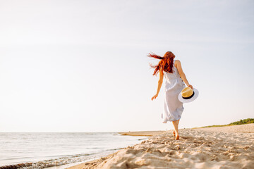 Happy young woman in dress and straw hat and walking alone on empty sand beach at sunset sea shore and smiling. Freedoom, vacation