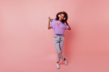 Wall Mural - Full-length portrait of amazing african lady in jeans holding skateboard. Elegant black girl in headphones and purple shirt standing on pink background.