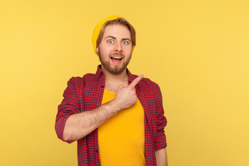 Look, advertise here! Surprised excited hipster guy in beanie hat looking at camera with happy amazed expression, pointing away at copy space, blank yellow background for commercial idea. isolated