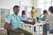 © Seventyfour - Portrait of young African-American man looking at camera while sitting at table during meeting with business team in background, copy space
