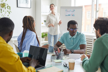 Wall Mural - Portrait of multi-ethnic business team discussing project while sitting at table in conference room with manager standing by whiteboard in background, copy space