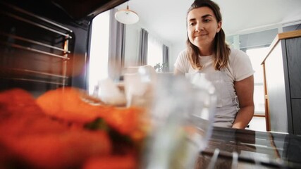 Wall Mural - food cooking, culinary and people concept - young woman with potholder taking baking dish with salmon fish and vegetables out of oven at home kitchen