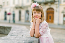 Little Beautiful Blonde Girl With Pink Flower In Her Hair Wearing Pink Dress Posing Outdoor On Sunny Summer Autumn Day In Old City Lviv, Ukraine. City Outdoor Portrait Of Little Girl