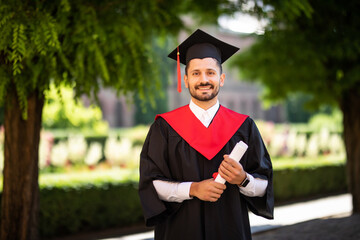 Wall Mural - Young male student dressed in black graduation gown. Campus as a background. Boy cheerfully smiling, holding diploma and looking at camera