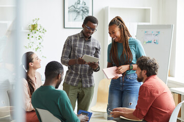 Wall Mural - Multi-ethnic group of people sitting in circle while discussing business project in office, focus on smiling African-American woman talking to colleague standing up , copy space