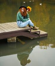Lonely Young Woman Standing On Lake Water Background. Redhead Girl Looks At Camera.  Water Surface Of The Lake. Reflection In Water Of Autumn Forest, Yellow Leaves In Hand.