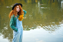 Lonely Young Woman Standing On Lake Water Background. Redhead Girl Looks At Camera.  Water Surface Of The Lake. Reflection In Water Of Autumn Forest, Yellow Leaves In Pocket.
